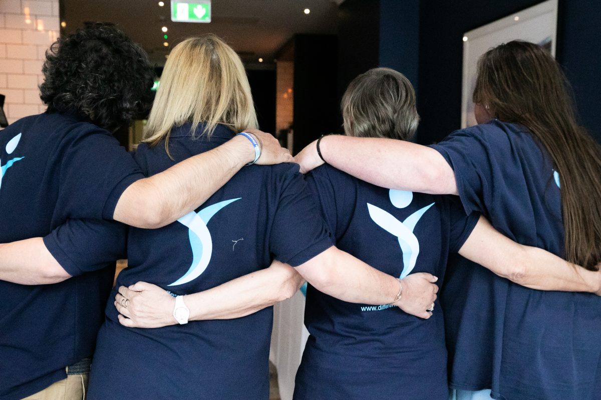 Four Different Strokes volunteers with arms around each other's shoulders, seen from behind, wearing navy Different Strokes t-shirts
